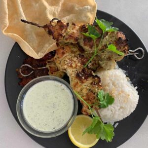 Chicken malai boti skewers with rice, pappadum, mint yoghurt and lemon on a black plate