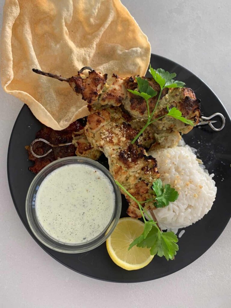 Chicken malai boti skewers with rice, pappadum, mint yoghurt and lemon on a black plate