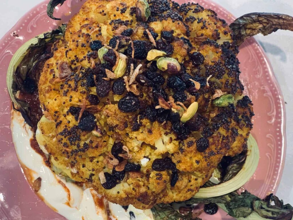 Whole roasted Indian spiced cauliflower topped with currants, pistachios and mustard seeds on yoghurt