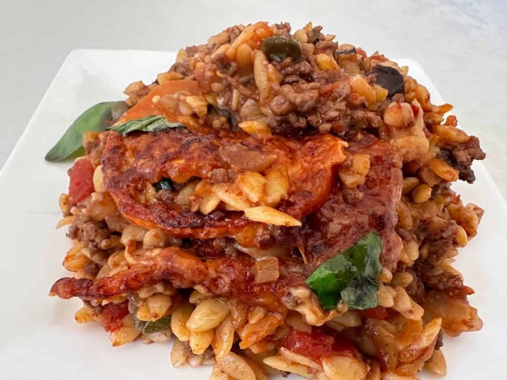 Baked puttanesca pasta with beef, olives, melted cheese and fresh basil on a white plate