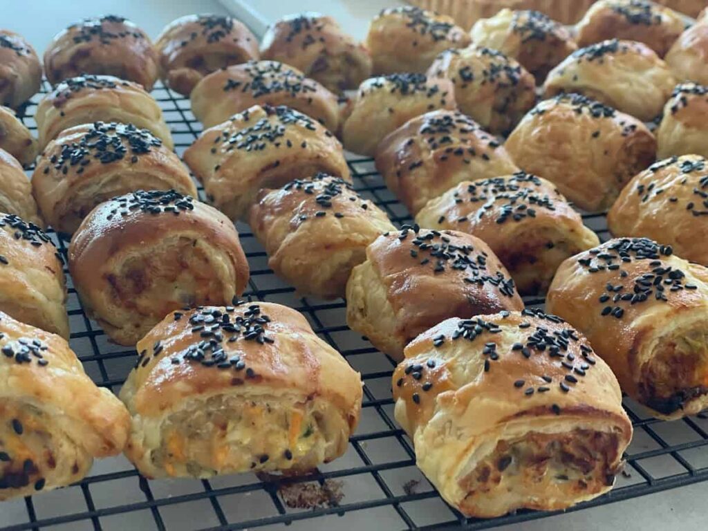 Golden homemade sausage rolls topped with black sesame seeds cooling on a wire rack
