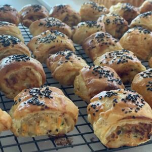 Golden homemade sausage rolls topped with black sesame seeds cooling on a wire rack